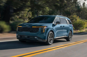 A blue 2025 Kia Carnival driving down a single lane road next to a wooded area near Orangeburg, South Carolina